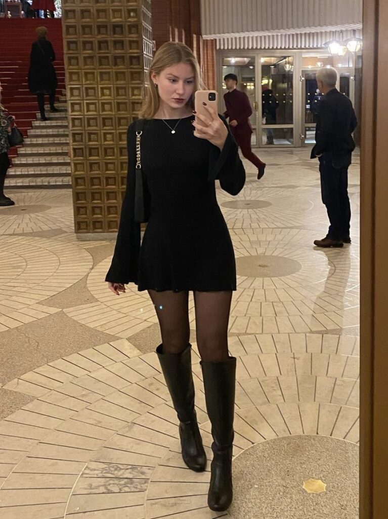 Woman taking a mirror selfie in a black dress with knee-high black boots and a stylish black purse, perfect for a night out.