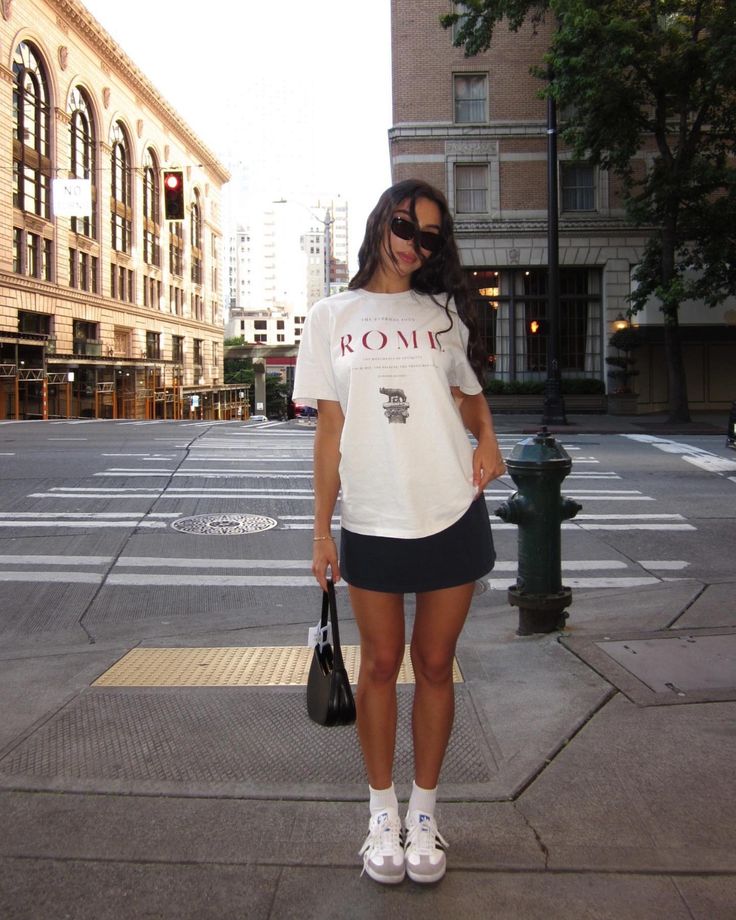 Woman in a casual street style outfit with a graphic tee, mini skirt, and white sneakers, standing on a city sidewalk.