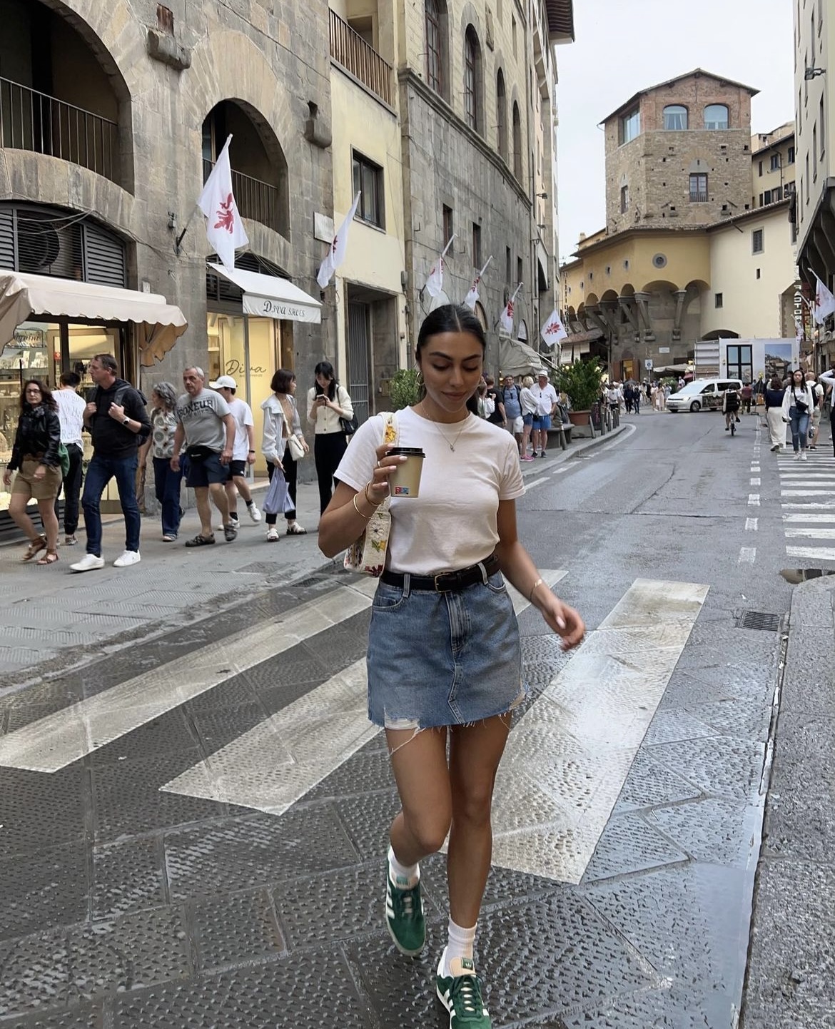Woman walking on a cobblestone street in a chic, casual outfit with a denim skirt, white tee, and green sneakers, holding a drink.