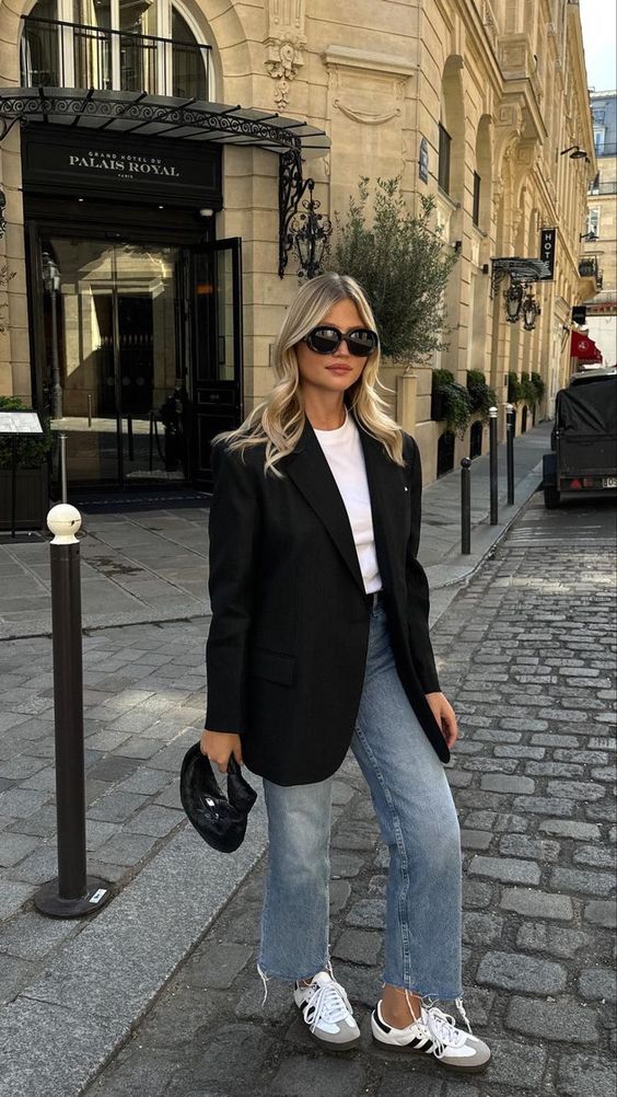 Woman in chic street style with oversized black blazer and light-wash jeans standing in front of Palais Royal hotel entrance.