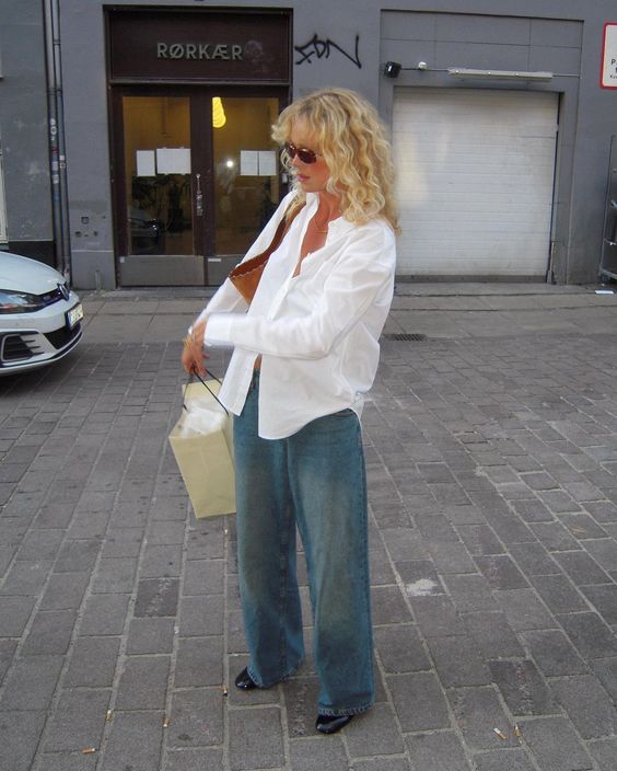 Woman on a city street with wide-leg jeans, a white blouse, and beige shoulder bag.