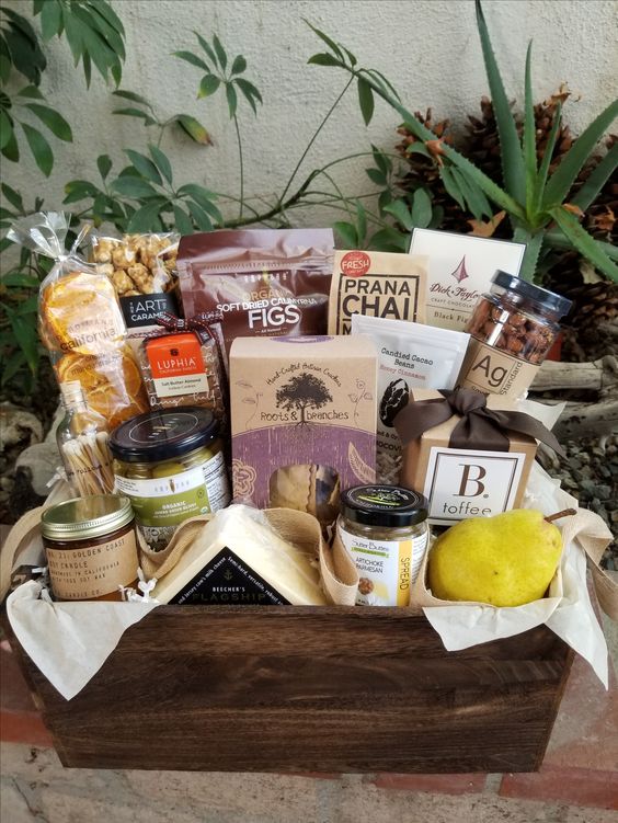 A gourmet food basket with an assortment of treats such as dried figs, chai mix, and toffee, presented in a wooden crate with a lush plant background.