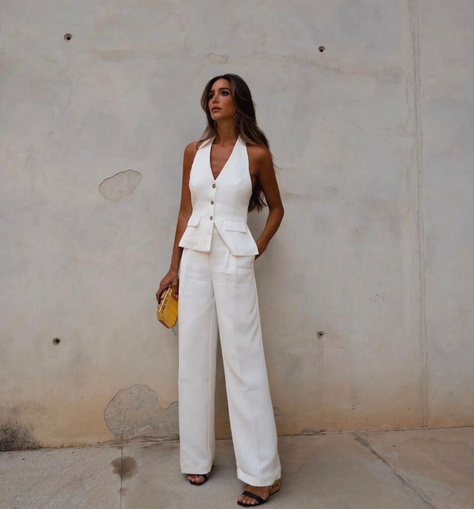 A woman stands against a neutral-toned wall with subtle textures. She wears a chic white ensemble, featuring a sleeveless, peplum top with a V-neckline and matching wide-leg trousers. Her accessories include black open-toe sandals and a small yellow clutch. She gazes off to the side with her hair styled straight, cascading over her shoulders.