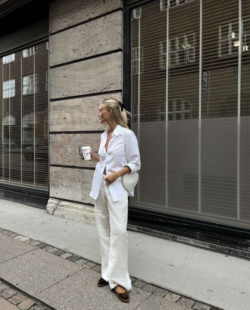 A woman in a relaxed white outfit stands on a city sidewalk. She is wearing an unbuttoned white shirt paired with linen trousers, complemented by dark loafers. She holds a coffee cup in one hand and carries a white handbag with the other. Her blonde hair is styled back into a ponytail with a casual black hair tie, and she appears to be smiling while looking to her right. Behind her, there’s an urban backdrop featuring a concrete building with large windows covered by brown blinds.