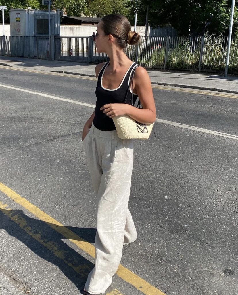 A woman crosses an urban street on a sunny day, wearing a casual summer outfit. She has on a black tank top with contrasting white trim, paired with light cream-colored linen trousers. Her hair is styled into a neat bun at the back of her head, and she sports dark sunglasses. Over her shoulder, she carries a textured cream-colored shoulder bag. She appears to be walking with a purpose, her gaze directed towards something out of frame.