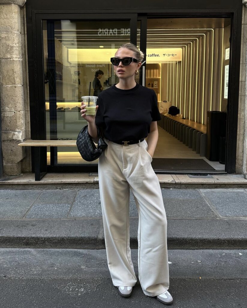 A woman stands on a city sidewalk, holding a cup of iced coffee. She is wearing a chic black t-shirt neatly tucked into high-waisted cream trousers, paired with white toe-cap flats. Her look is accessorized with large black sunglasses, gold hoop earrings, and her hair is pulled back into a sleek bun. She carries a black textured shoulder bag. The backdrop features a modern storefront with reflective windows and a deep interior.