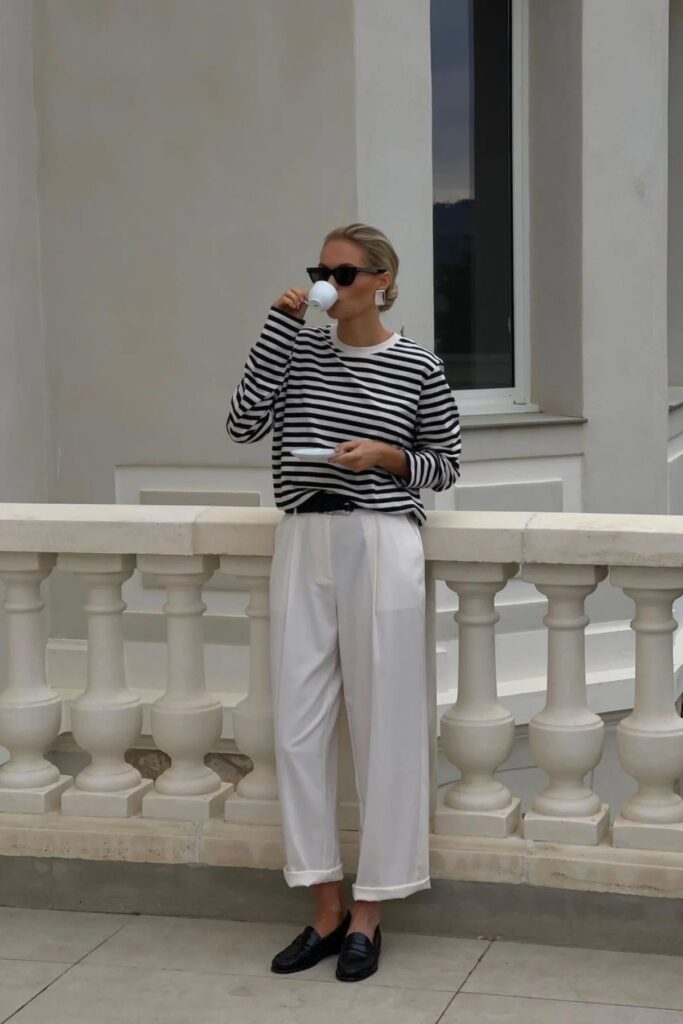 A woman enjoys a serene moment on a balcony with classic balustrades. She sports a timeless look in a black and white striped long-sleeve shirt, tucked into high-waisted white trousers, and finishes the outfit with black loafers. She sips from a white cup, her other hand holding the saucer, and wears sleek, large sunglasses. Her hair is pulled back into a low bun, and she accessorizes with hoop earrings. The scene exudes a relaxed, elegant vibe.
