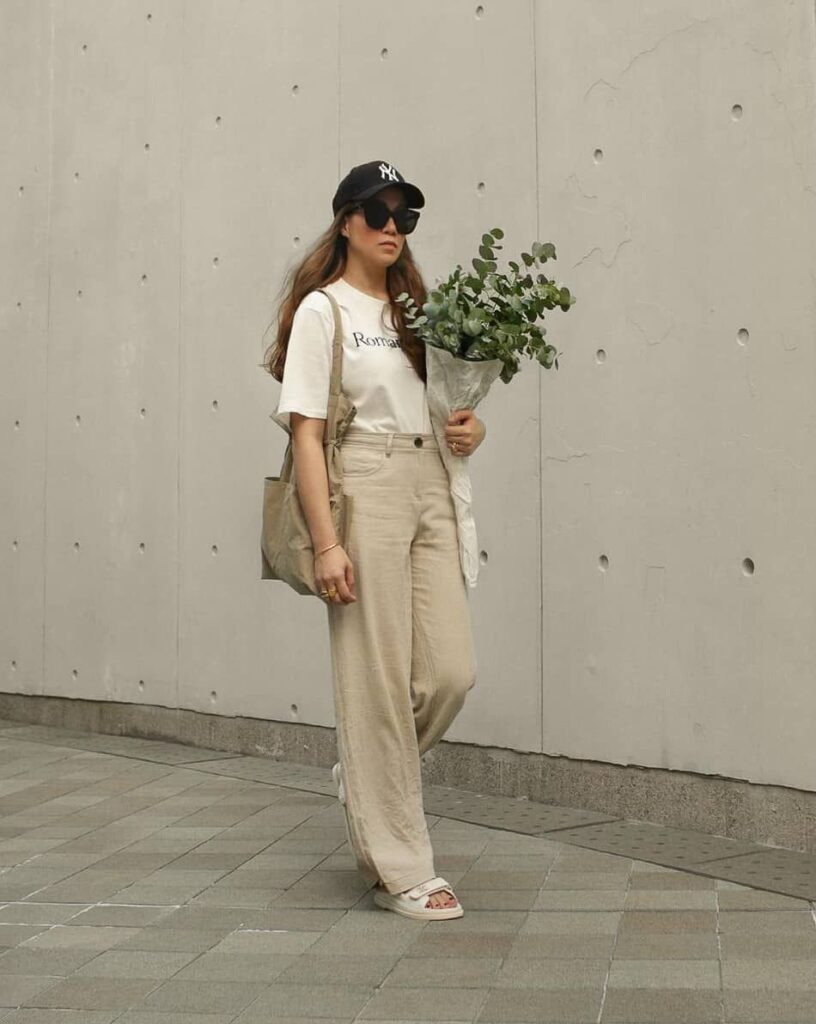 A woman in a casual chic outfit walks along a paved area against a minimalist concrete wall. She wears a white t-shirt with a print, light beige pants, and white slide sandals. Her look is accessorized with a black baseball cap, dark sunglasses, and carries a canvas tote bag. She holds a bouquet of eucalyptus leaves wrapped in paper, adding a touch of green to the neutral tones of her ensemble. Her posture and stride suggest movement and purpose.
