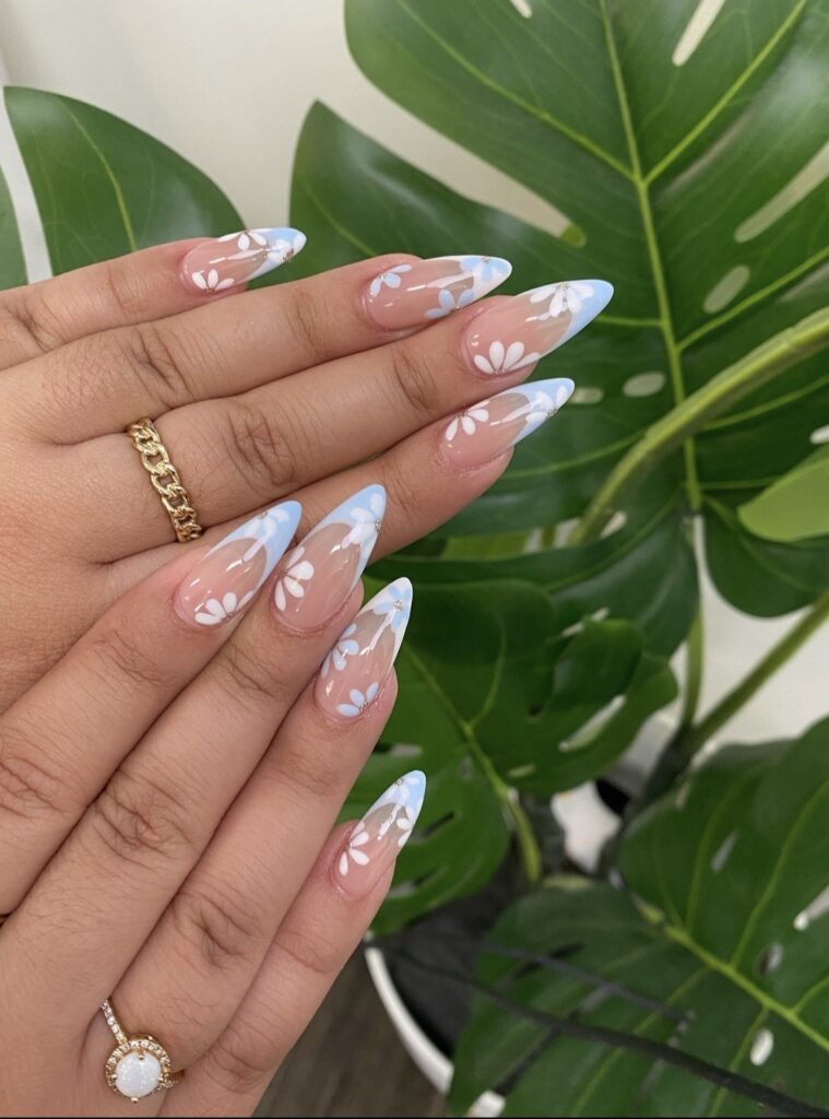 Almond-shaped nails with a translucent base, sky-blue French tips with white floral accents, and inverse design on ring fingers featuring white tips with blue flowers.