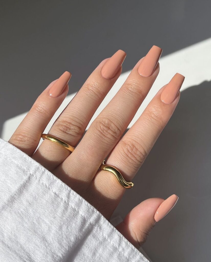 A photo displaying a hand with nails polished in a soft, peachy hue, reminiscent of a peaceful summer dusk, adorned with elegant gold rings.