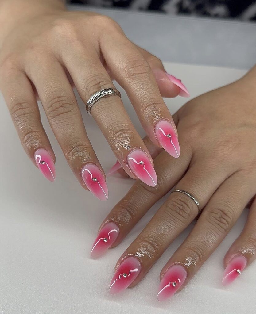 A hand displays stiletto nails with an ombre effect that transitions from a vibrant strawberry pink to a soft, milky pink, each nail crowned with a small sparkling rhinestone.