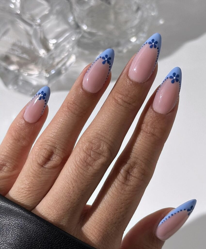 A hand shows off nails with a delicate pink base and charming blue daisy designs on the tips, a fresh and floral nail art perfect for the sunny season.