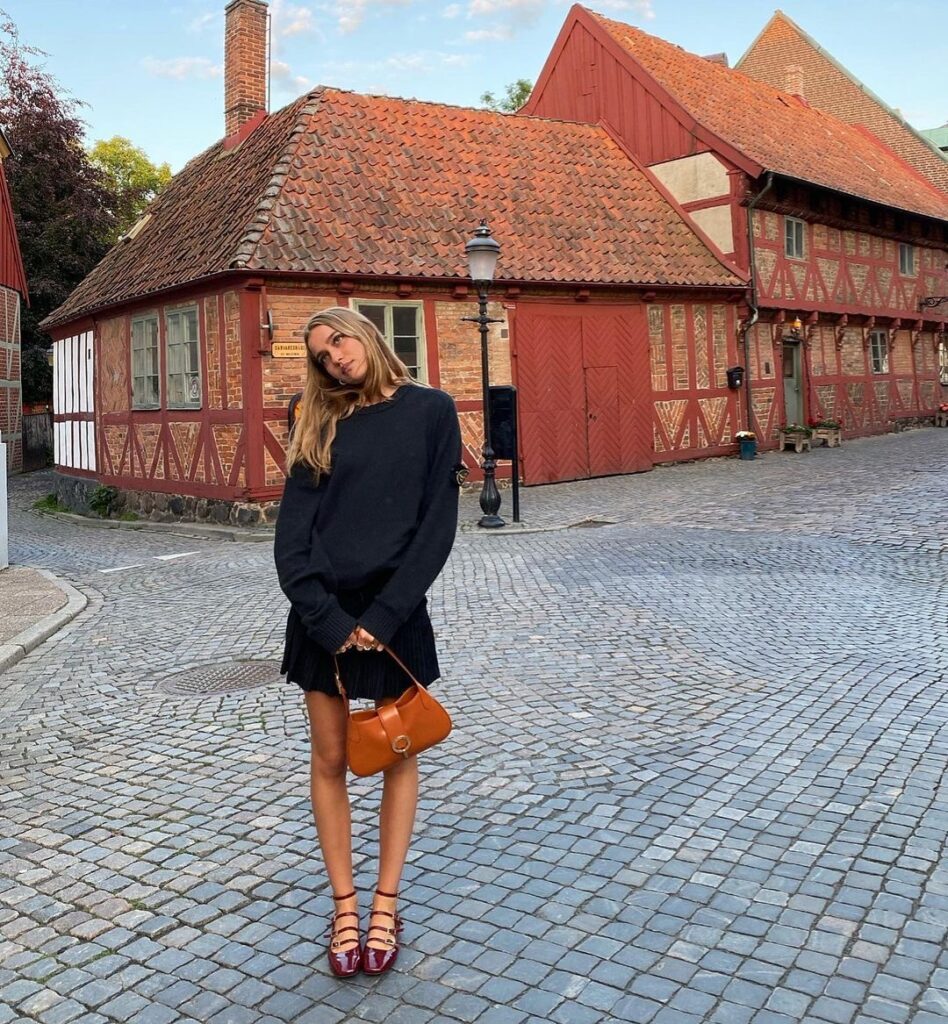A woman poses on a cobblestone street, pairing a navy sweater with a pleated mini skirt and burgundy strappy ballet flats, holding a dark beige shoulder bag.