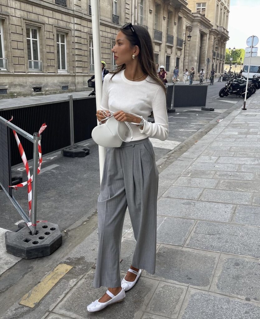 A casual chic outfit displayed by a woman wearing grey trousers and a long-sleeve white top, complemented with white ballet flats, standing on a Parisian street.