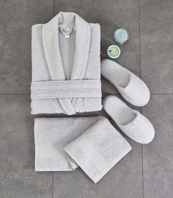 A gray bathroom set comprising of a soft bathrobe, matching slippers, and towels with a candle on a stone surface.