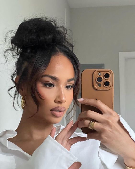 A woman with a stylish high bun hairstyle, featuring loose pieces framing her face, holds a smartphone for a mirror selfie. She wears a white blouse, showcasing a chic and elegant look.