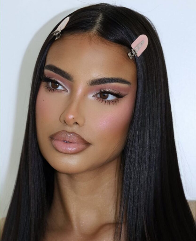 This look features expertly contoured cheeks that create a sharp, defined appearance, paired with perfectly groomed bold eyebrows. Neutral pink matte eyeshadows are complemented by a subtle winged eyeliner, while the lips are coated in a glossy beige lipstick. The look is completed with a sleek, straight hairstyle and chic hair clips for a polished finish.