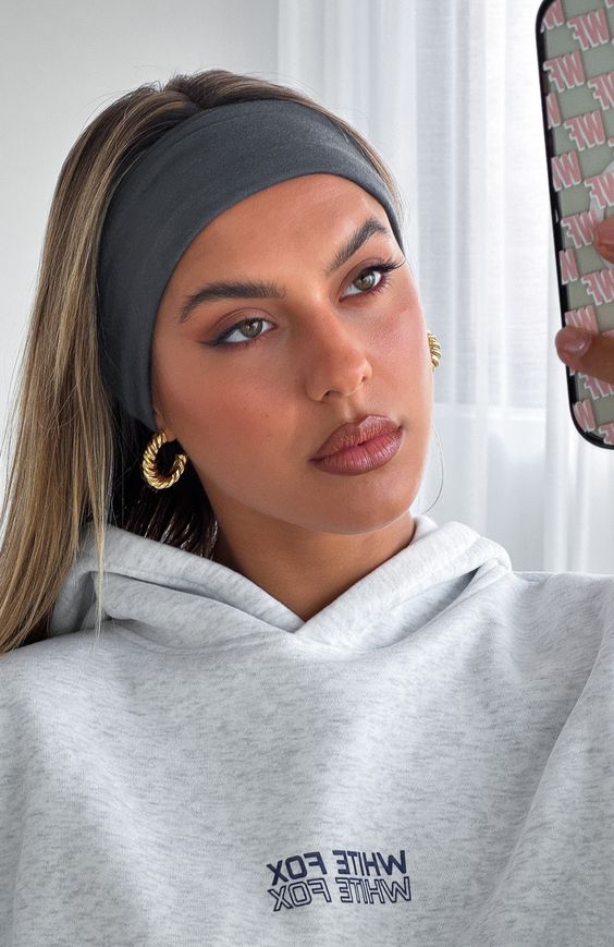 A woman in a casual setting wearing a gray hoodie and a gray headband, taking a selfie. Her makeup is natural, complemented by bold hoop earrings, highlighting a relaxed yet fashionable style.