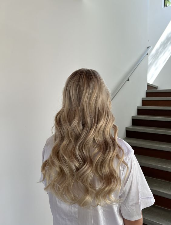 A woman standing on a staircase, her back to the camera, displaying long, flowing hair styled in soft, natural waves. She wears a simple white shirt, emphasizing a relaxed yet stylish vibe.