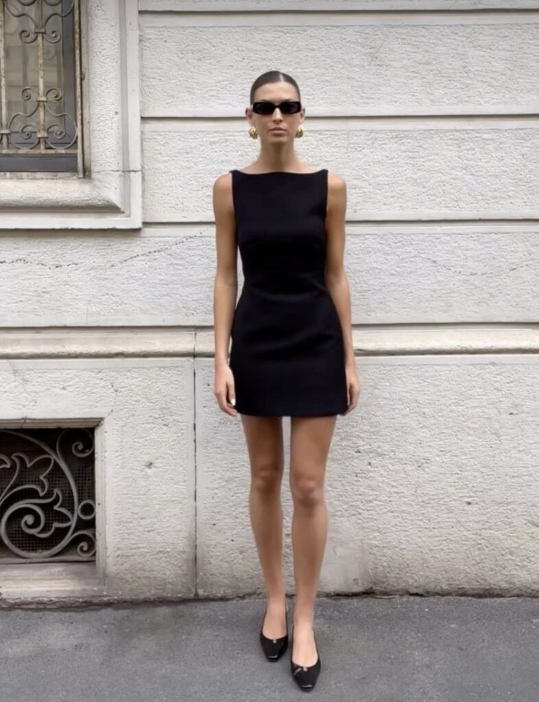 Woman standing against a white wall, wearing minimal jewelry, black ballet flats, and sunglasses for a chic minimalist look.