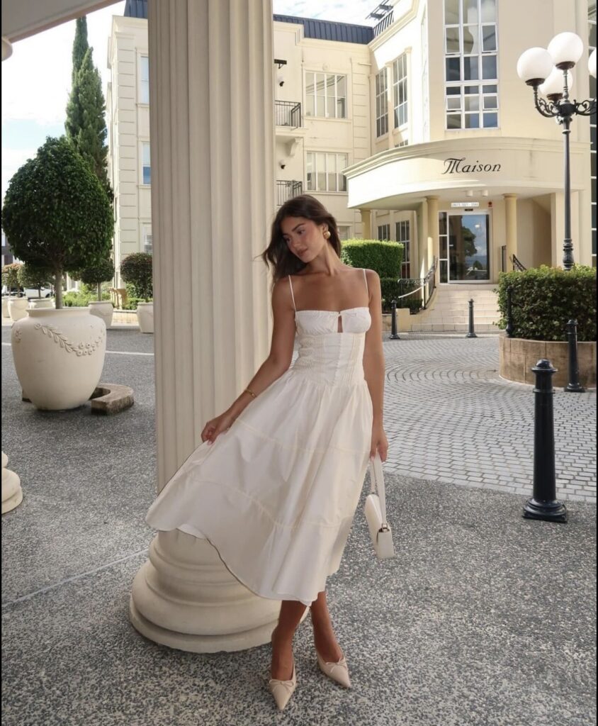 A woman in a white midi dress with spaghetti straps, posing elegantly in an outdoor setting with classical architecture, including tall columns and neatly trimmed bushes.