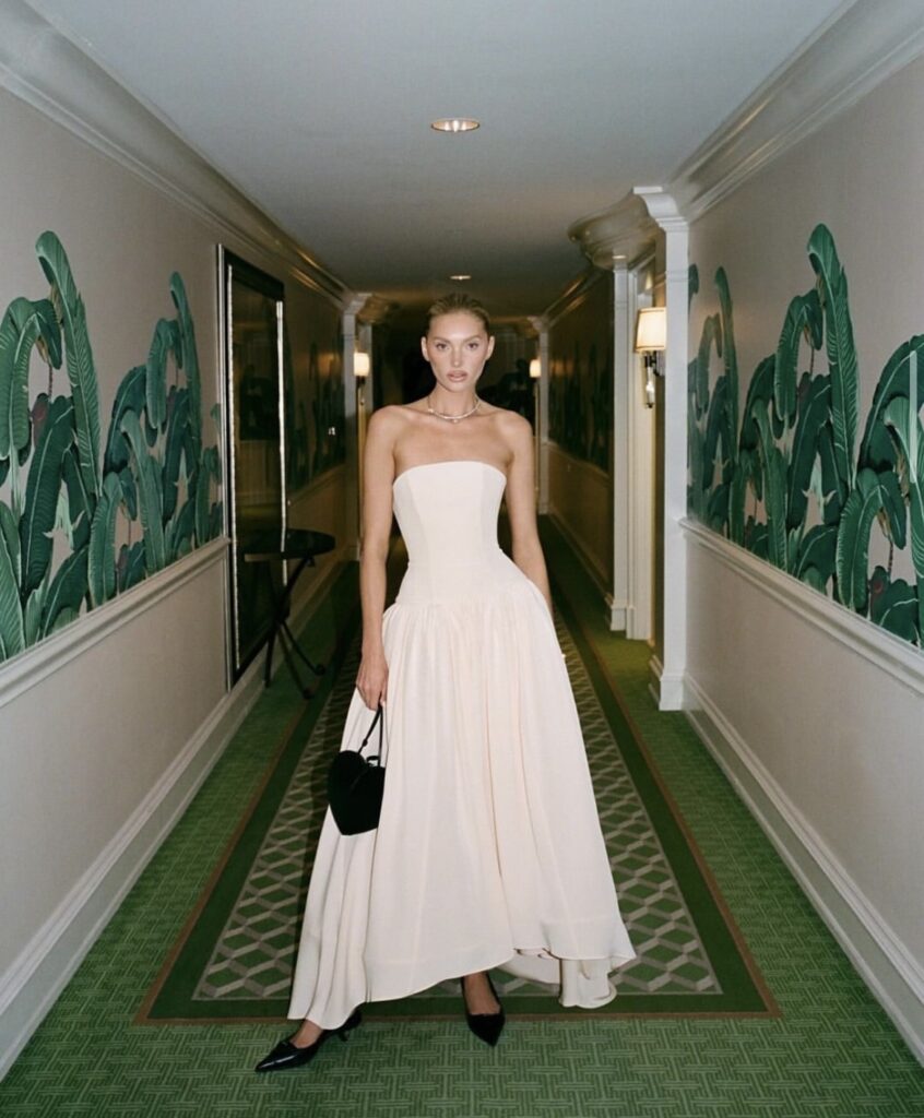 A woman in a strapless, floor-length white gown, holding a small black handbag, standing in a hallway with patterned green carpet and banana leaf wallpaper.