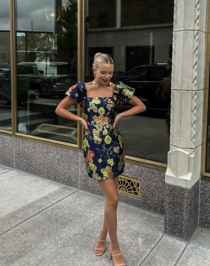 A woman in a short, dark blue dress with bold floral prints in yellow and red, featuring puff sleeves, posing confidently on a city sidewalk.