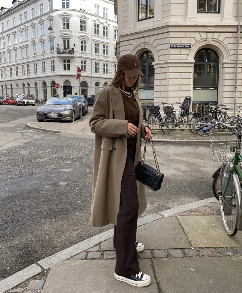 Brown wide-leg pants paired with a brown sweater and a light brown trench coat, accessorized with a black handbag and casual sneakers.