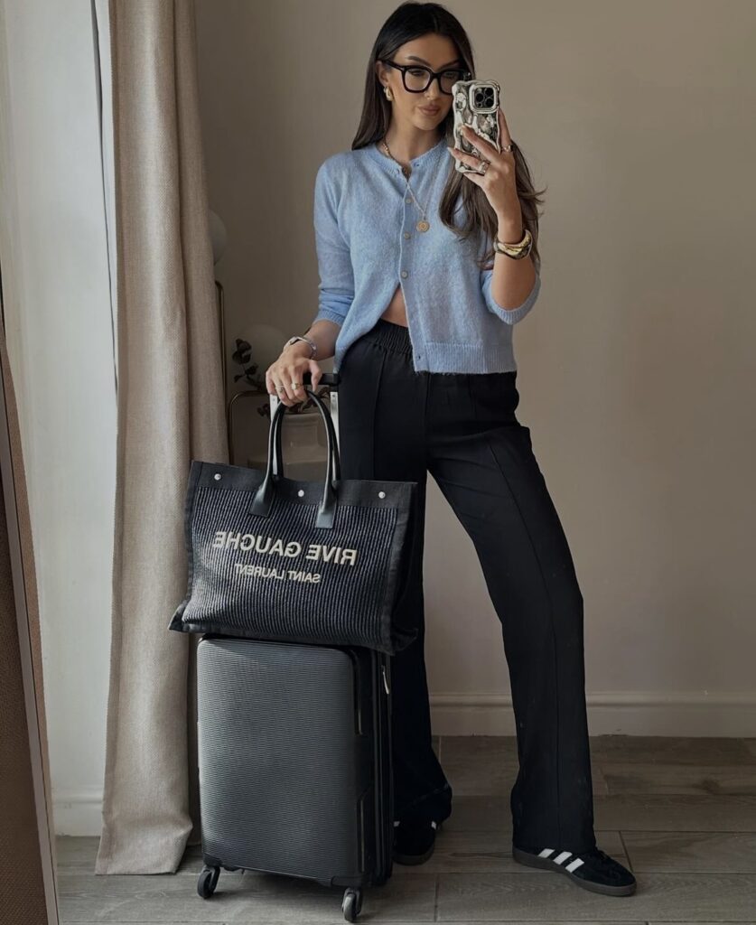 A stylish travel outfit featuring a light blue cardigan, black relaxed-fit trousers, and white sneakers, paired with a structured tote bag.