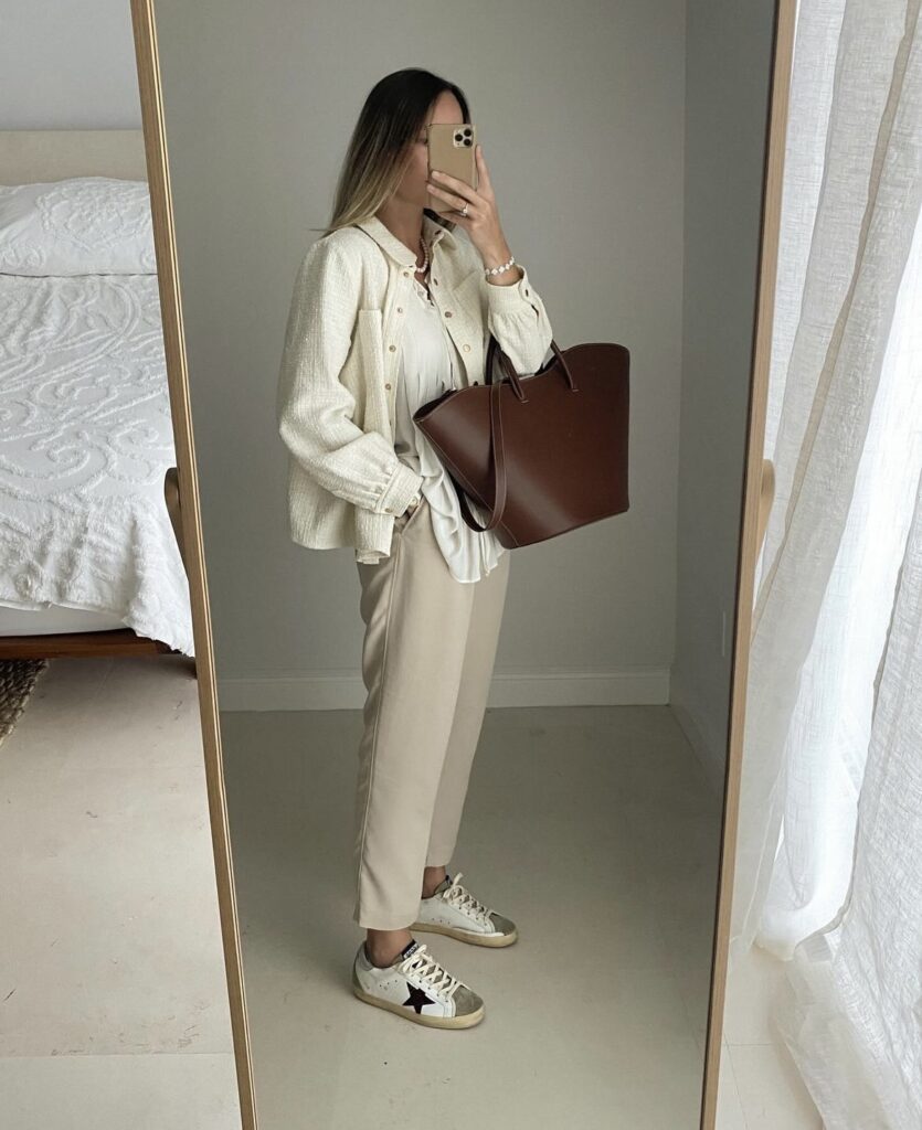 An elegant travel outfit with a cream-colored cardigan, flowy white blouse, beige trousers, and white sneakers, completed with a large brown tote bag.