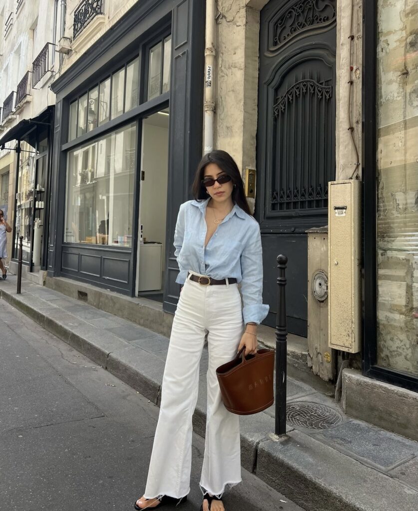 Woman wearing a blue button-down shirt and white pants with black sandals and a brown handbag.