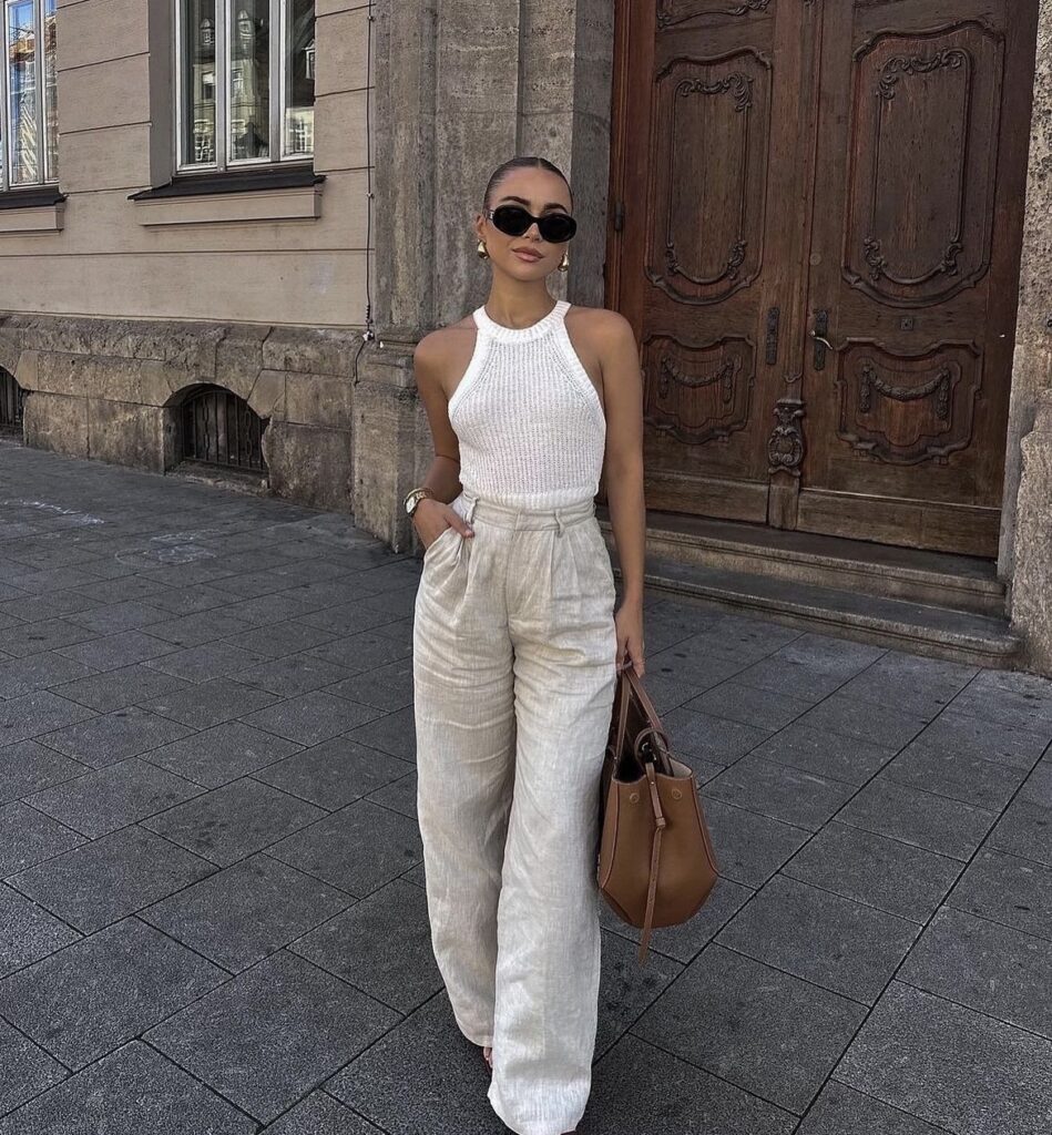 Woman in a knit top and linen pants with flat sandals and a straw bag.