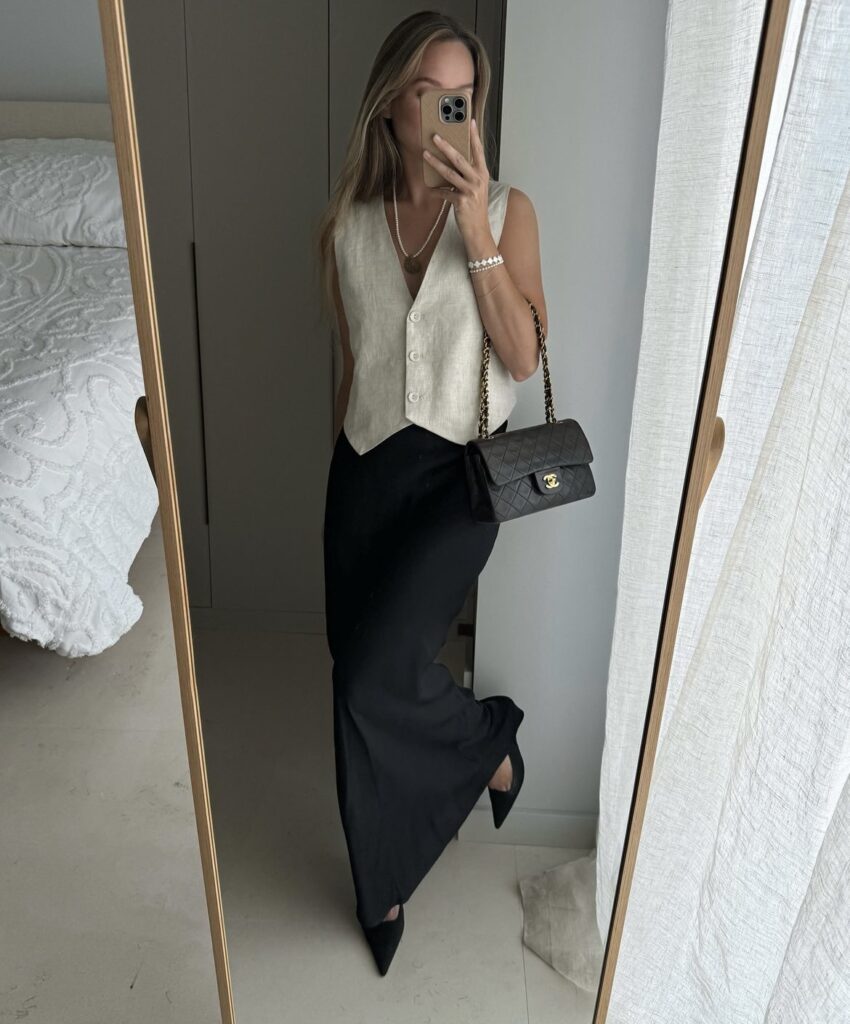 Woman wearing a vest and black maxi skirt with black flats and a black handbag.