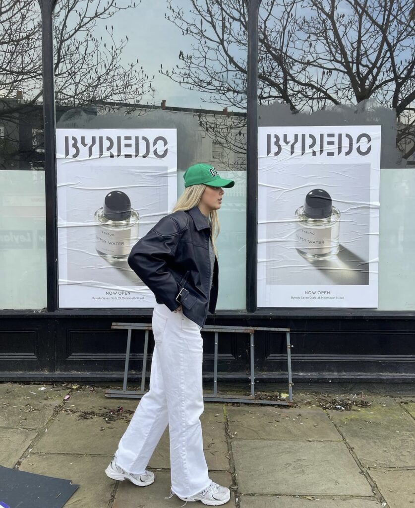 Woman in a leather jacket and white jeans with white sneakers and a green baseball cap.