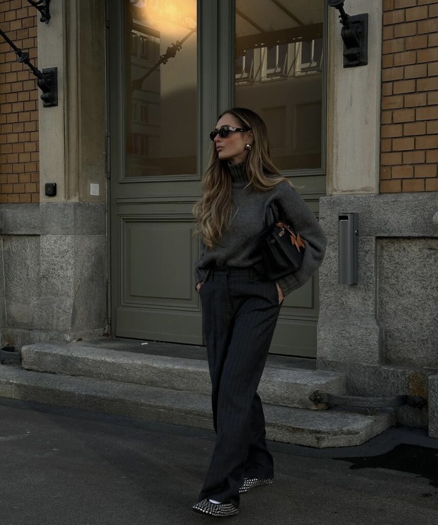 Woman wearing a grey turtleneck sweater with dark grey trousers and a black handbag.