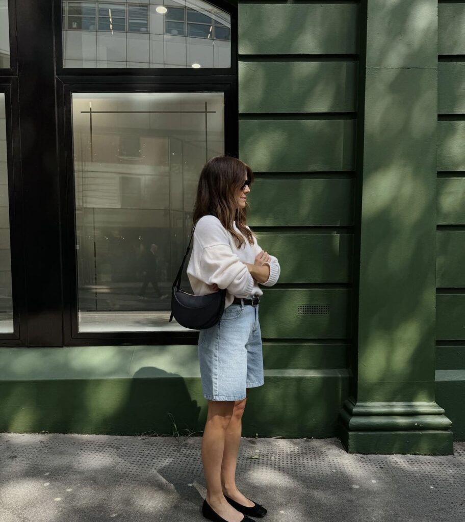 Woman wearing a cozy sweater with high-waisted denim shorts and black ballet flats.