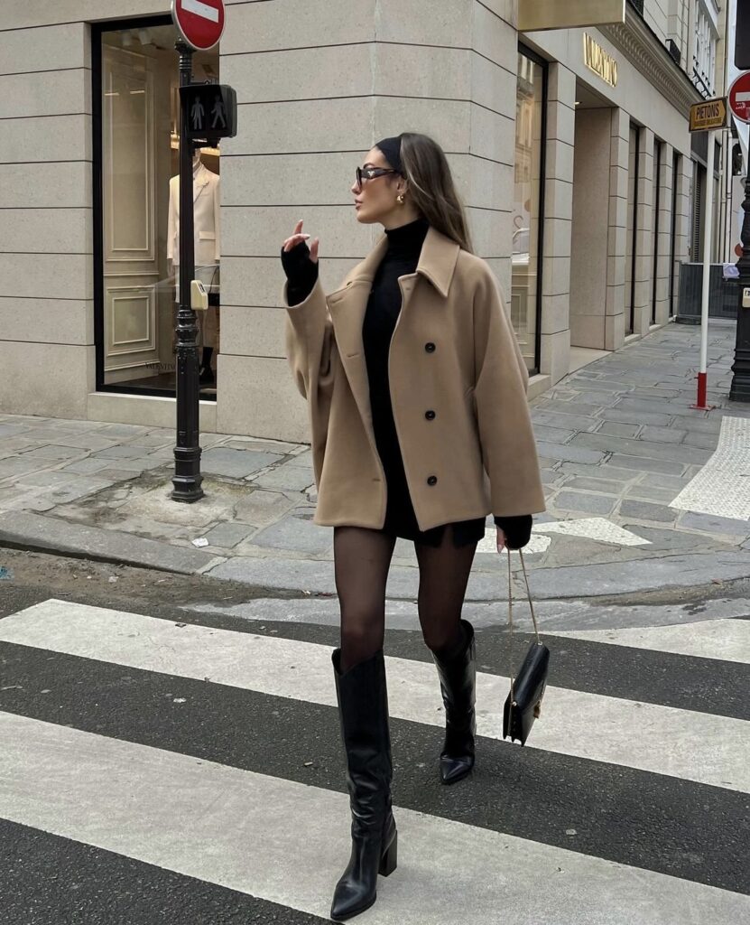 A camel coat over a black dress with knee-high boots and a minimalist handbag.