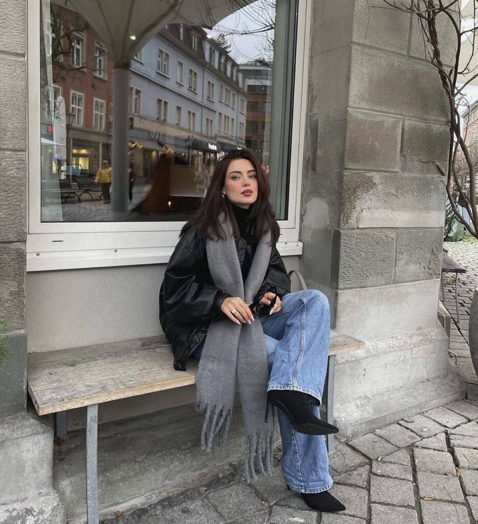 An oversized black leather jacket with blue jeans, a grey scarf, and boots.