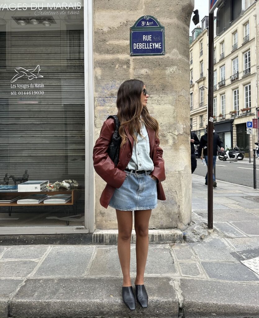 A woman wearing a bold red jacket, paired with a denim mini skirt. The outfit is completed with oversized sunglasses and a structured black handbag.