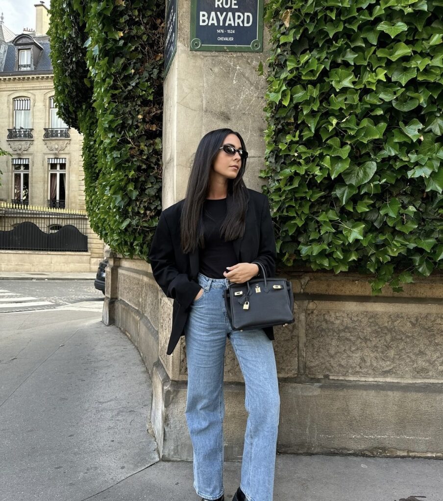 A woman is wearing high-waisted jeans paired with a classic black blazer. She completes the outfit with a black handbag.