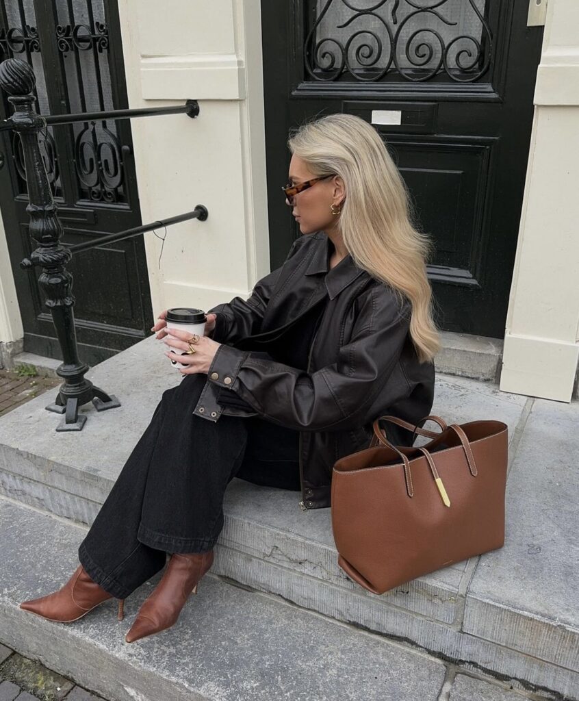 A woman wears a brown leather jacket over black jeans. The look is finished with pointed-toe brown boots and a large brown tote bag.