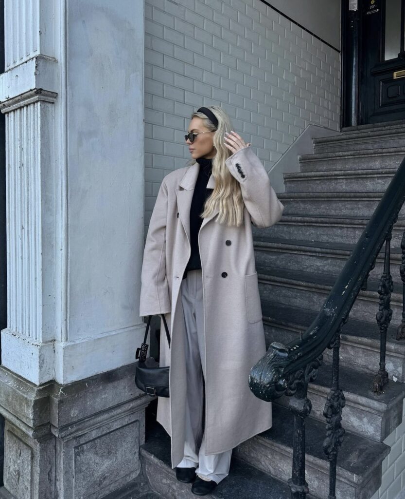 A woman is dressed in a classic trench coat, paired with wide-leg trousers and a black turtleneck. She accessorizes with a small black handbag and sunglasses.