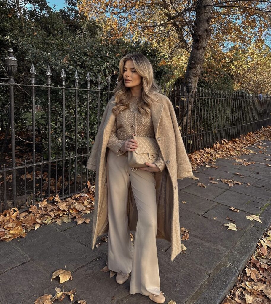 A woman showcases an all-beige ensemble featuring a knitted cardigan and beige trousers, layered with a long beige coat. The look is completed with neutral ballet flats and a quilted handbag.