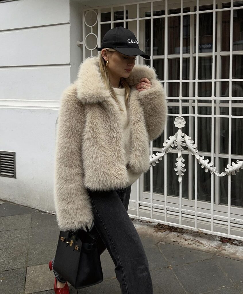 A woman is wrapped in a light beige faux fur jacket, paired with black jeans and a cozy knit sweater. She tops off the look with a black baseball cap.