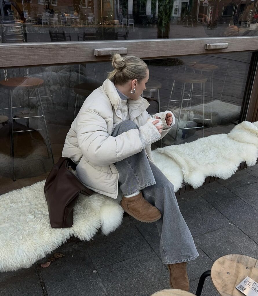 A woman is wearing a cream-colored puffer jacket, paired with grey jeans and brown boots.