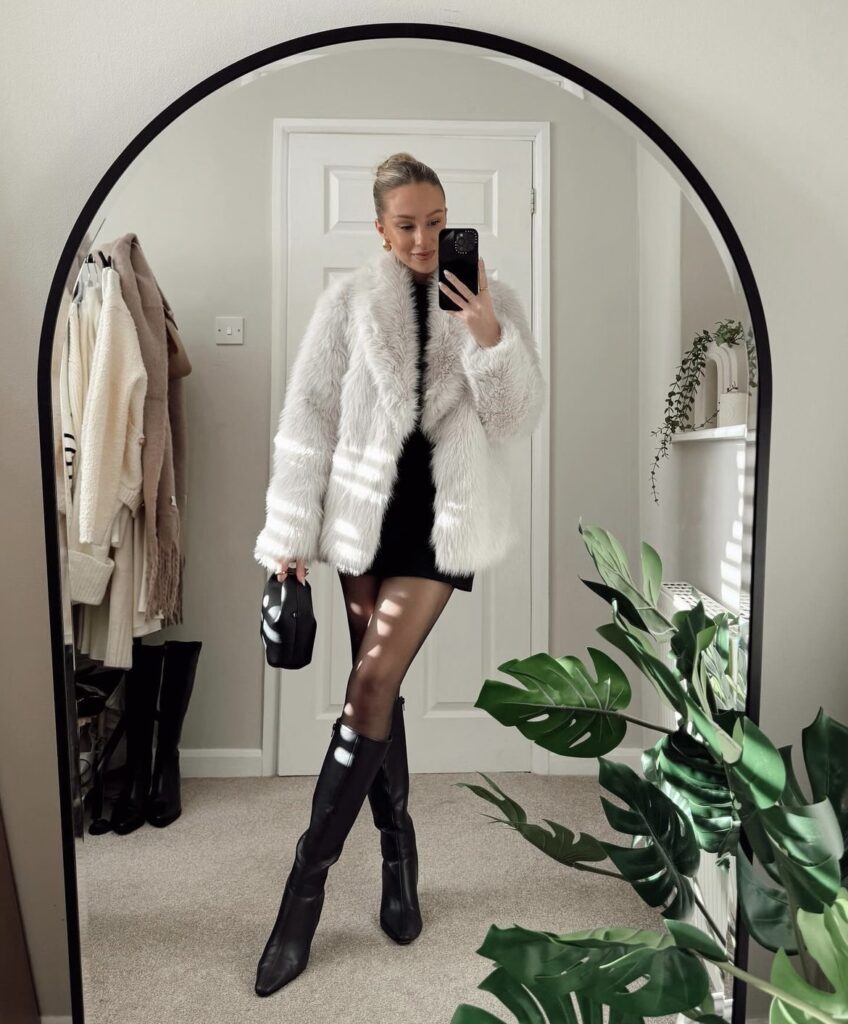 A stylish winter outfit featuring black knee-high boots, a black mini dress, and a white faux fur jacket. The look is completed with sheer tights and a small black handbag.