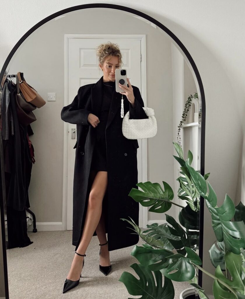 Woman in a black dress styled with a long black coat, paired with black pointed-toe heels and holding a white handbag.