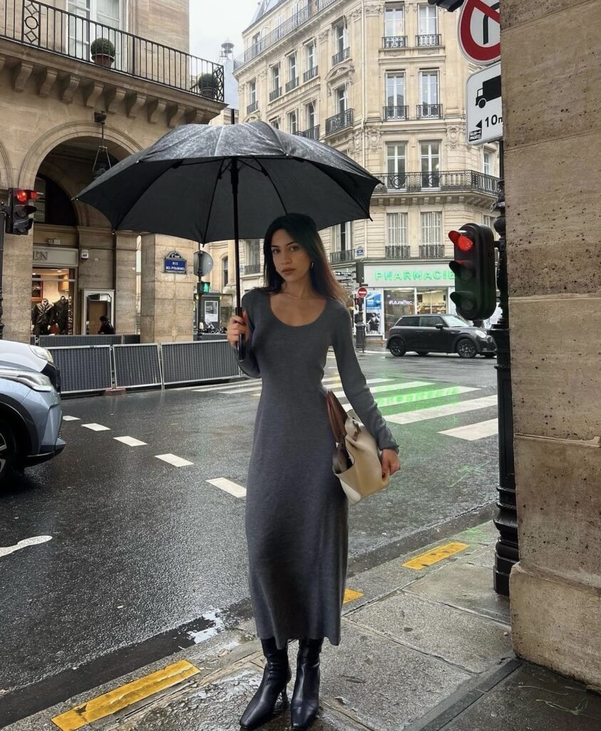 Woman wearing a grey maxi dress paired with black boots, holding an umbrella on a rainy day in the city.