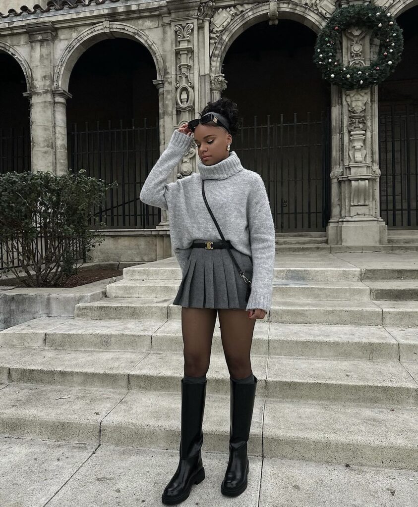 Black knee-high boots styled with a grey pleated skirt and a matching turtleneck sweater, finished with sheer tights and a black crossbody bag for a soft, monochromatic winter look.