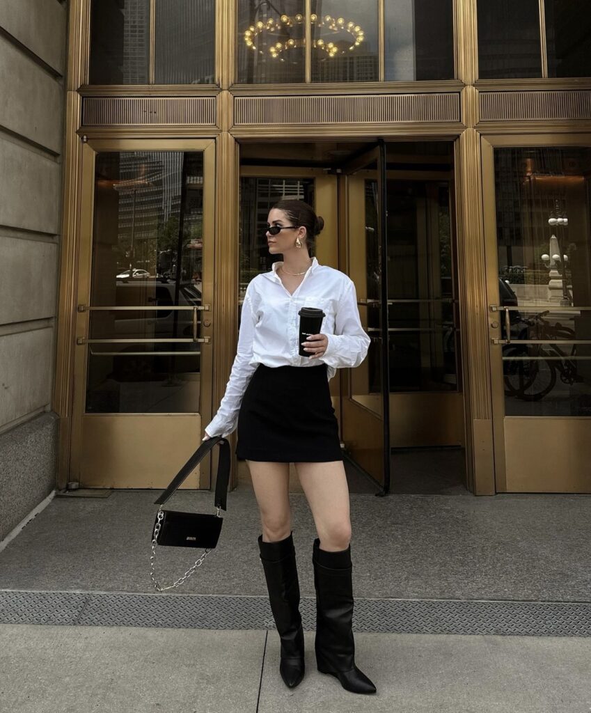 A chic winter outfit featuring black knee-high boots, a black mini skirt, and a crisp white button-down shirt, accessorized with a black structured handbag and sunglasses.